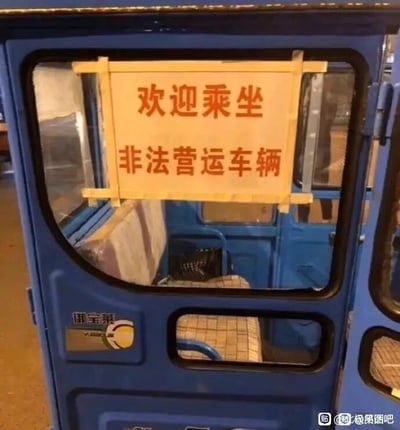 这是一张原始拍摄的静态单图，主体为一辆蓝色的“御宝莱”牌电动三轮车。在车厢的侧窗位置，用黄色胶带固定着一块白底红字的长方形牌子，牌子上清晰地写着“欢迎乘坐 非法营运车辆”。这种直白到毫不掩饰的标语和常规营运车辆会刻意规避“非法营运”表述的做法形成强烈反差，制造出滑稽搞笑的效果。车厢内部可见简易的格纹座椅，车门上印有“御宝莱”的品牌标识，图片右下角带有相关水印。