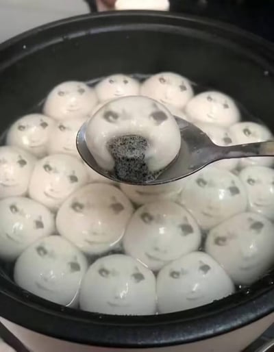图中展示了一锅正在被煮的汤圆，所有汤圆的表面都被P上了同款的人脸，人脸带着微妙又诡异的微笑。其中一个汤圆被盛放在勺子里，露出了内部的黑芝麻馅，原本普通温馨的煮汤圆场景，因为这些人脸的加入变得十分猎奇，充满了搞笑与诡异交织的反差感，让人觉得既滑稽又有点不适。