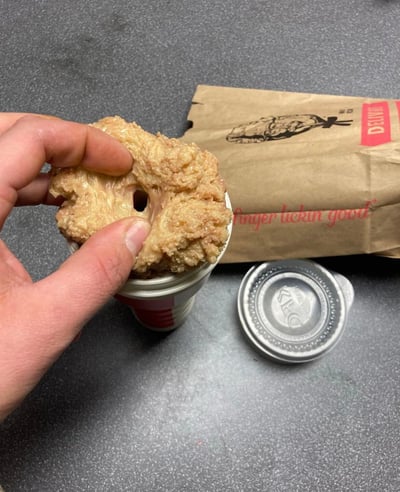 一只手正捏着一块造型奇特的KFC炸鸡，这块炸鸡意外呈现出甜甜圈般的环形结构，中间有一个明显的圆孔，外皮金黄酥脆，纹理清晰。炸鸡下方是一个KFC的白色纸杯，旁边散落着印有上校桑德斯头像和经典标语“finger lickin good”的棕色纸袋，以及一个银色的KFC饮料杯盖。整体画面展示了一次有趣的快餐体验，因食物的特殊形状产生了喜剧效果。