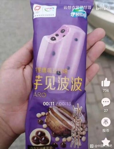 这是一段B站视频的截图，画面中一只手拿着伊利牧场的芋见波波冰淇淋，包装为紫色主色调，上方印有冰淇淋本体的效果图，是芋泥紫色搭配黑色波波球的样式，包装标注了“芋泥鹰嘴豆口味”“芋见波波”“冰淇淋”等字样，下方还有冰淇淋内馅的展示图。画面右侧是B站的互动按钮区域，显示点赞736、评论27，视频进度条显示当前播放至11秒，总时长13秒。