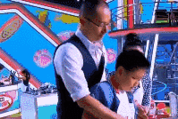 这是一段来自综艺《超级育儿师》的GIF动图，画面里一位身着白衬衫搭配黑背心的戴眼镜成年男性，从身后环抱住穿蓝红拼色上衣的小男孩，二人处于色彩鲜亮的综艺录制场地的操作台前，似乎在进行手工或烹饪类的引导教学，整体场景偏向亲子教育类的综艺环节，没有双关、谐音梗等内容。