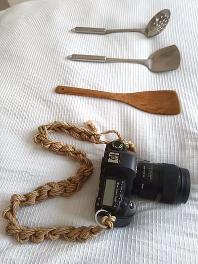 一张静物摄影照片，展示了在白色纹理背景上精心排列的物品组合。画面中有一台黑色单反相机，配有编织绳材质的相机带，旁边整齐摆放着三件厨房用具：两把金属勺子（其中一把带孔洞）和一把木质锅铲。物品布局呈现出从大到小、从近到远的层次感，相机位于前景位置最为醒目，厨房用具则在背景依次排列。整体风格简约自然，可能用于生活方式分享或物品展示。