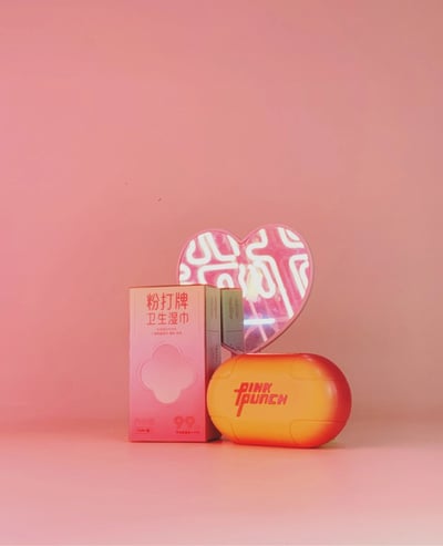 粉色渐变背景上展示了三款产品：左侧是粉打牌卫生湿巾的粉色包装盒，中间是带有迷宫图案的粉色心形镜子，前方是橙黄色渐变的PINKY PUNCH椭圆形产品。整体构图简洁，色彩以粉色和橙色为主，营造出甜美活泼的视觉效果。