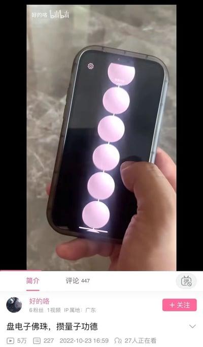 图片展示一只手握着智能手机，手机屏幕上垂直排列着多个粉色圆形图标，模拟电子佛珠的交互界面。底部显示Bilibili视频平台的界面元素，包括上传者“好的咯”的信息（6粉丝、1视频、IP属地广东）、视频标题“盘电子佛珠，攒量子功德”，以及5万播放量、227点赞、发布时间2022-10-23 16:59和27人正在观看的实时数据。
图片通过“盘电子佛珠”“攒量子功德”的创意，将传统宗教文化中的“盘佛珠积功德”行为与现代科技概念“电子”“量子”结合，形成幽默的反差感，调侃了科技对传统习惯的趣味改造。