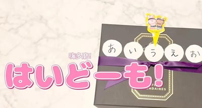 图片展示了一个放置在白色大理石台面上的黑色礼品盒，盒身系有紫色丝带，顶部装饰着一个黄色卡通钥匙扣，钥匙扣上有两只并排站立的兔子形象。礼盒正面贴有五个圆形白色标签，分别写有日语五十音图中的「あ、い、う、え、お」。图片左侧有醒目的粉色日文文字「はいどーも!」及其中文谐音「嗨多磨!」，整体呈现出温馨可爱的日式礼品氛围。