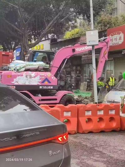 这是一处城市街边的施工场景，一台厦门远山机械的轮式挖掘机被改装成了亮眼的粉色，机身侧面印上了《魔卡少女樱》中的主角木之本樱的动漫形象，挖掘机顶部还堆放着几袋物品。画面近处有比亚迪秦PLUS汽车，周围设置了橙色施工围挡，背景是临街商铺，还有穿着反光背心的施工人员在现场。硬核的工程机械设备搭配软萌的二次元少女涂装，形成了强烈的反差感，十分吸睛。图中没有双关、谐音梗或暗示内容。