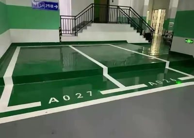 图片展示了一个室内停车场场景。地面被涂成绿色，带有白色线条划分出两个停车位，编号分别为“A024”和“A027”。背景中有一段带黑色栏杆的楼梯通向更高楼层，楼梯旁的墙壁上挂着一个蓝色指示牌，上面写着“2号楼”并配有向右的箭头。墙壁为绿色和白色相间，地面光滑有反光，整体环境整洁，未见人员或车辆。