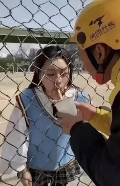 这是一张利用视觉错位效果拍摄的原始静态照片，场景位于户外的铁丝网旁。画面中，一位穿着蓝白学院风服装的女生站在铁丝网后方，嘴部贴近铁丝网；另一侧是身着美团外卖工作服、头戴黄色头盔的外卖员，手持一个白色杯子面向女生。通过铁丝网的网格，女生的嘴部和外卖员手中的杯子形成了视觉错位，看起来仿佛女生正在从杯子中饮用液体，实际二者并无接触，营造出有趣的互动效果。