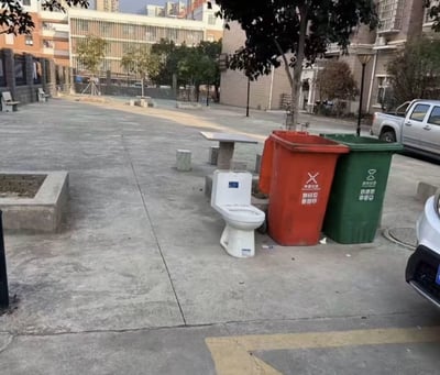 这是一张居民社区公共区域的实拍照片，画面右侧的垃圾分类投放点旁摆放着一个被住户丢弃的完整白色陶瓷坐便器，紧邻坐便器的是红色有害垃圾桶和绿色厨余垃圾桶；背景可见社区内的石制休闲桌椅、行道树、居民楼建筑，还有停放的皮卡车等车辆，呈现出住户丢弃废旧大件卫浴用品的现实场景，坐便器紧邻垃圾桶摆放的状态形成了荒诞的视觉效果。