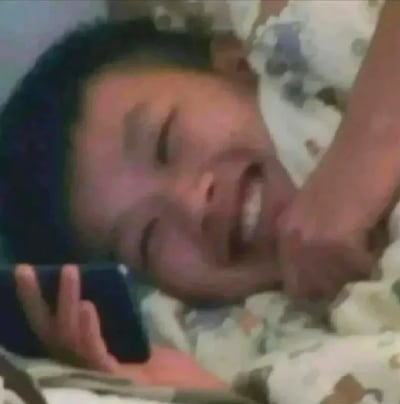 图片中显示一个人躺在床上，头枕在有花纹的枕头上，正在打电话。此人眼睛紧闭，嘴巴张开，露出牙齿，表情看起来非常开心，正在大笑。另一只手轻轻抚摸着打电话的人的脸颊。整个场景给人一种轻松愉快的感觉。