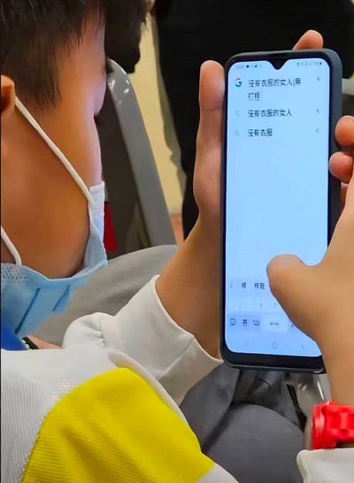 整张图为实拍竖图，画面主体是一名佩戴蓝色医用口罩的小男孩，他正坐在座位上操作智能手机，手机屏幕显示搜索引擎界面，男孩正在输入“没有衣服的女人”相关关键词，下方也出现了对应的联想搜索词；男孩身穿白黄拼色的上衣，手腕佩戴红色电子表，画面呈现出未成年人进行不符合其年龄的搜索行为的反差搞笑感。
