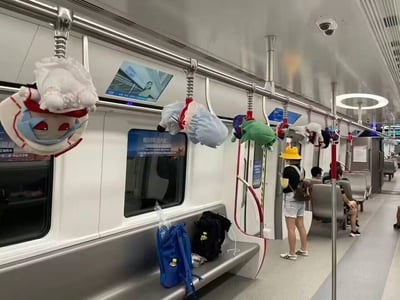 这是一张在地铁车厢内拍摄的真实场景图，原本的金属吊环把手被多个二次元动漫角色的毛绒玩偶替代，一个个玩偶挂在把手的弹簧下，成为了独特的“人形吊环”。车厢内有几位乘客，一位戴着黄色帽子的乘客站在车厢连接处，其他乘客坐在座位上，座位上还放置着背包等随身物品。车窗上贴有“保利海德天汇”的房地产商业广告，整体是日常的地铁通勤环境，挂满玩偶的设计充满了二次元爱好者的巧思，为单调的通勤空间增添了新奇有趣的氛围，没有双关或谐音梗内容。