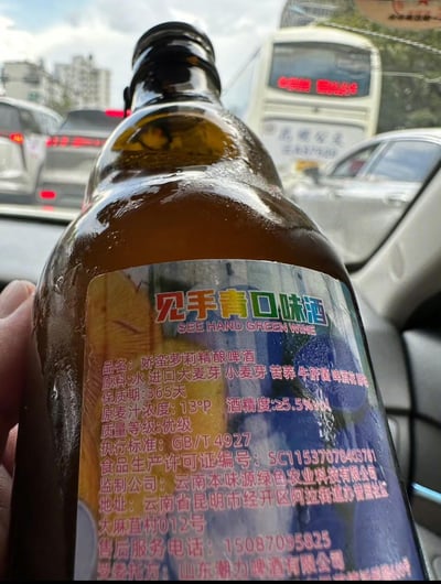 图片中一只手握着一瓶深棕色啤酒，瓶身表面有冷凝水珠，标签醒目印有“见手青口味酒”和“SEE HAND GREEN WINE”字样，下方标注实际品名为“娇蛮萝莉精酿啤酒”。标签详细列出原料（水、进口大麦芽、小麦芽、苦荞、牛肝菌、啤酒花等）、保质期365天、原麦汁浓度13°P、酒精度≥5.5%vol等信息，监制公司为云南本味源绿色农业科技有限公司，地址位于云南省昆明市经开区。背景为车内视角，可看到窗外行驶的车辆和公交车，整体场景像是在车内拍摄的产品实拍图。