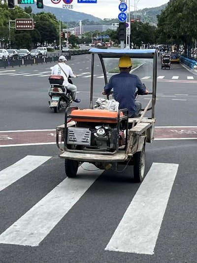 这是一张城市道路路口的实拍照片，画面前景是一辆由金属框架搭建的自制简易机动三轮车，车辆后部搭载着外露的发动机装置，驾驶员头戴黄色安全帽，身穿深蓝色带点上衣，正驾驶车辆处于斑马线上；画面左侧有一名身穿白色上衣的男子，正驾驶电动车在道路上行驶。背景可见城市道路的交通信号灯、指示牌、行道树，远处还有山峦和其他车辆，整体呈现出日常城市交通的场景，这辆结构简单、发动机外露的自制三轮车是画面的特殊看点，推测是用于户外作业的改装车辆。