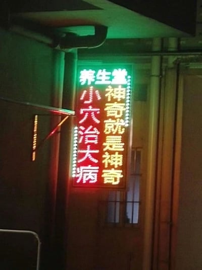 图片展示了一个挂在室内墙面的立式霓虹灯招牌，整体呈竖长条形，边框有白色灯珠装饰。招牌顶部以绿色发光字体显示“养生堂”，下方分两列用黄色发光字体书写“小穴治大病”和“神奇就是神奇”，背景为红色。招牌周围可见金属管道和昏暗的环境光线，推测为小型养生或中医理疗店铺的宣传标识。招牌文字直白强调穴位疗法的效果，具有民间店铺常见的夸张宣传风格。