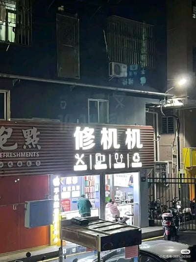 这是一张夜晚拍摄的城市居民区街边照片，画面核心是一家名为“修机机”的手机维修店。店铺招牌设计玩了谐音拆字的趣味梗：将“修手机”的概念拆解成“亻| 乚 | 乚 | 木”的视觉符号，对应“修机机”的文字，用拆字的方式呼应“修手机”的业务，让招牌更具记忆点。店内有两位顾客，一位身着绿色上衣站在柜台前，另一位粉色上衣的顾客坐着，似乎在等待维修服务或进行咨询。店铺左侧是一家名为“包點”（附带英文“REFRESHMENTS”）的点心店，店门为红色。画面右下角可见停放的电动车和汽车，背景是带有防护栏、空调外机的居民楼，楼体在夜色中显得昏暗，整体场景充满了城市居民区夜晚的生活气息，展现了街边小店的日常风貌。