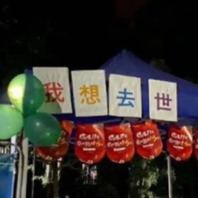 这是一张夜晚户外场景的实拍图，画面主体是蓝色帐篷上方悬挂的四块白色方形标语牌，分别以红、黄、蓝、棕四种颜色的字体写着“我”“想”“去”“世”，组合成字面带有消极情绪的语句；标语牌下方挂着一排红色的日式风格灯笼，灯笼上印有白色的艺术字体，具体内容模糊不清；画面左侧还点缀着三颗绿色的气球。这是典型的年轻人整活设计，通过看似消极的反差标语吸引路人注意力，大概率是校园社团招新或者线下趣味活动的引流手段，用夸张的反向表达制造搞笑效果，并无真正的消极倾向。