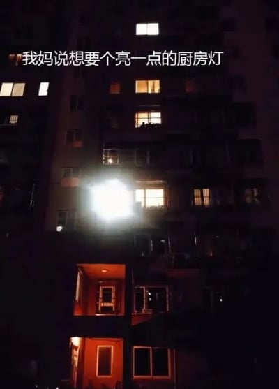 这张图片是夜晚拍摄的居民住宅楼实拍图，整栋楼处于夜色中，只有零星住户的窗户透出正常亮度的居家灯光，其中某一户的厨房位置安装的灯亮度极高，在楼外拍摄都能看到强烈的过曝白光，光线格外醒目。图片上方配文“我妈说想要个亮一点的厨房灯”，通过配文和实际灯光亮度的夸张反差制造笑点，调侃为了满足长辈想要亮厨房灯的需求，最终安装的灯具亮度过高，超出正常使用范畴的搞笑情况。