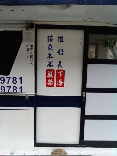 这是一张拍摄民用船只舱门区域的照片，舱门及旁边的船身印有各类文字信息：舱门上的竖排标语因排版问题产生了搞笑的断句歧义。右侧竖排蓝色文字为“推船長”，下方搭配红底白字“下海”；左侧竖排蓝色文字为“搭乘本船”，下方搭配红底白字“嚴禁”。按照中文竖排从右到左、从上到下的阅读顺序，很容易被误读为“推船長下海，搭乘本船嚴禁”，意思变成“把船长推下海，严禁搭乘这艘船”，但结合船身标注的“乘客人數20人”“船員4人”的载客信息来看，这显然不符合船只的运营逻辑，正确的标语意图应该是严禁推船长下海以及搭乘本船的相关规定，却因排版失误形成了极具反差感的搞笑歧义。此外，船身还印有重复的数字“9781”，右上角有一个圆形的“釣魚 文化休閒”标识。
