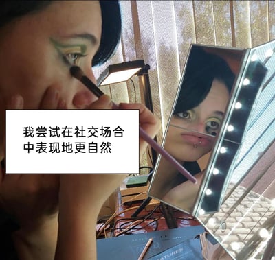 这是一张原创实拍的静态图，画面中一位带有唇钉的人正对着带LED灯的三面化妆镜，手持化妆刷专注地绘制着绿黄配色的夸张眼妆，镜子里清晰呈现出她的倒影。图中添加了文字标注“我尝试在社交场合中表现地更自然”，形成强烈的反差感：用极具个性的夸张妆容来试图让自己在社交场合中更自然，这种反差带有自嘲意味，大概率是想通过打造独特的妆容来建立自信，以此在社交场景中更放松自在。