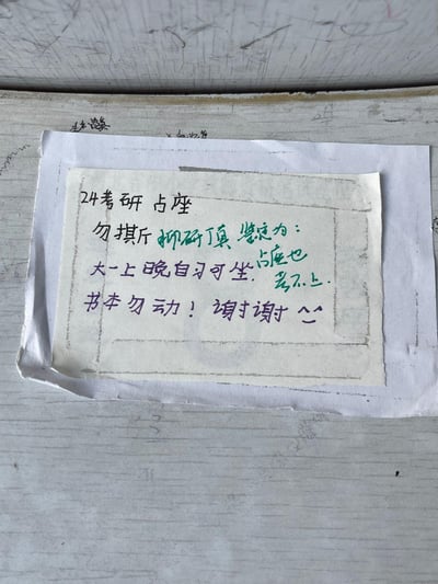 这是一张贴在高校自习室桌面的手写占座纸条，为24考研的学生所写。纸条中用谐音梗“抑研真”对应“考验真”，自嘲“占座也考不上”，既表明了占座需求，又带着幽默的自嘲感；同时贴心留出座位给大一上晚自习的同学使用，最后提醒他人不要挪动自己的书本，态度友好又不失风趣，是校园考研日常里很真实的小细节。