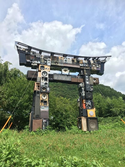 一张户外艺术装置照片，展示了一个由大量废弃音响、扬声器和功放等音频设备堆砌而成的拱门结构。这个创意拱门呈现出传统日式鸟居的造型，由各种尺寸、颜色和年代的音响设备拼接组合而成，包括黑色、棕色、米色等不同色调的箱体和喇叭单元。装置位于一片绿色山林前，背景是蓝天白云，周围有茂密的树木和草地，营造出自然与工业废弃品结合的艺术氛围。整个结构看起来相当庞大，底部用绳索进行了固定，展现了变废为宝的环保艺术理念。