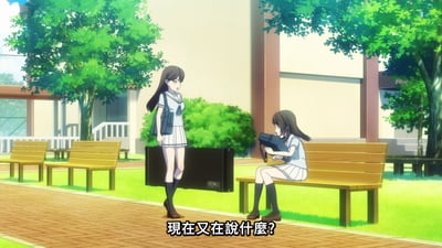 动画《MyGO!》中的校园场景，画面中有两位穿着蓝白校服的少女。一位站立在铺着红砖的小路上，身旁放着一个黑色的乐器箱，表情略带疑惑地看着另一位坐在木质长椅上的少女；坐着的少女则低头专注地抱着一个黑色物品。画面下方有字幕：“現在又在說什麼?”，营造出日常互动中的小疑惑氛围。背景有绿色的树木和教学楼，整体色调明亮清新。