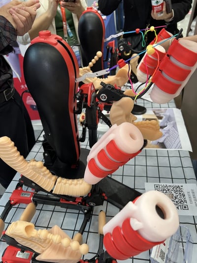 这张照片展示了在某个展览会上展出的机械康复设备。画面中央是一组黑色和红色相间的机械装置，包含多个关节和活动部件，看起来像是某种康复训练设备或假肢原型。装置上有米色的仿真肢体部分和彩色的连接线。背景中可以看到几位参观者正在观看展品，有人举起手机可能在拍照，还有人手持印有中文标识的饮料瓶。桌子上有一张带有QR码的纸张，可能用于获取更多产品信息。整个场景呈现出科技展览的氛围，展示了医疗康复领域的创新产品。