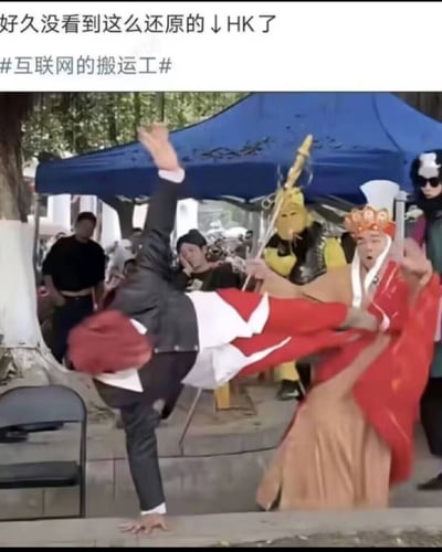 图片展示了一个户外场景，一名穿西装的男子正在表演倒立，另一名身着传统服饰的人站在他面前，似乎在与其互动。背景中可以看到蓝色帐篷和一些围观的人群，营造出一种热闹的氛围。