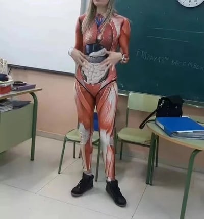这张静态图片拍摄于一间教室中，画面主体是一位女性教师，她穿着印有完整人体肌肉组织与腹部内脏器官图案的紧身连体衣，双手放在自己的腹部位置，似乎正借助这件特殊的衣服向学生直观展示人体内部结构。背景里能看到教室的桌椅、一块写有“12月13日 星期五”的黑板，还有挂在墙上的时钟，整体环境是典型的课堂场景，这种创意穿搭显然是为了让人体解剖相关的教学内容更生动、直观，帮助学生更好地理解知识。