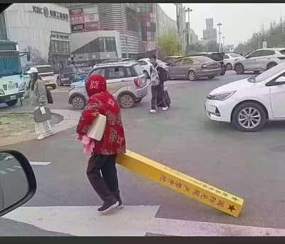 这是一张在城市路口拍摄的视觉错位照片，画面中一位穿着红色花棉袄的老人正走在过马路的途中，她身后的黄色国防光缆警示杆因为拍摄角度巧合，看起来像是从老人的身体里穿过去，制造出荒诞又搞笑的视觉错觉。背景里能看到其他行人、行驶的车辆，还有中国工商银行的建筑，是日常街头的一幕，却因为错位效果变得十分有趣。