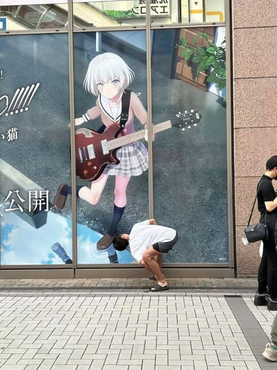 这是一张在日本街头拍摄的趣味错位照片：玻璃外墙上贴着《BanG Dream! It's MyGO!!!!!》里的角色要乐奈（白发持电吉他的女高中生形象）的宣传海报，一名穿白T恤的路人弯腰将头贴在海报中角色的脚边位置，利用平面海报与真实空间的视觉错位，制造出“被动漫角色踩在脚下”的搞笑效果，旁边还有一位戴口罩的黑衣路人路过。
无谐音梗或双关，仅通过错位构图制造趣味。