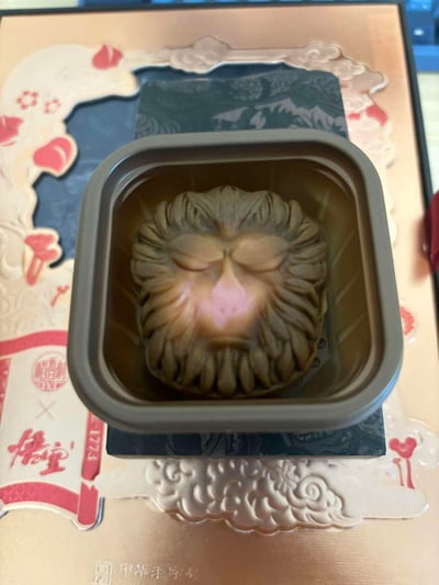 图片展示了一款造型精致的猴头月饼，放置在方形金属盒中。月饼表面雕刻有类似孙悟空的面部特征，双眼闭合，毛发纹理清晰，鼻部呈粉红色。包装盒采用传统中式设计，背景有云纹和红色花卉图案，左下角可见「儒堂」字样印章和「LZ73」标识。整体呈现出浓厚的传统文化氛围与创意食品设计的结合。