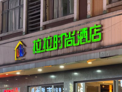 图片展示了一栋建筑的外立面，上方有一个醒目的霓虹灯招牌，主体文字为绿色的“拉拉时尚酒店”，左侧配有一个彩色的房屋形状logo，内部有“lala”字样。建筑外墙为浅棕色条状瓷砖，上方有几扇带窗帘的窗户，招牌下方有一排照明灯光，整体呈现出夜晚或傍晚时分的街景氛围。