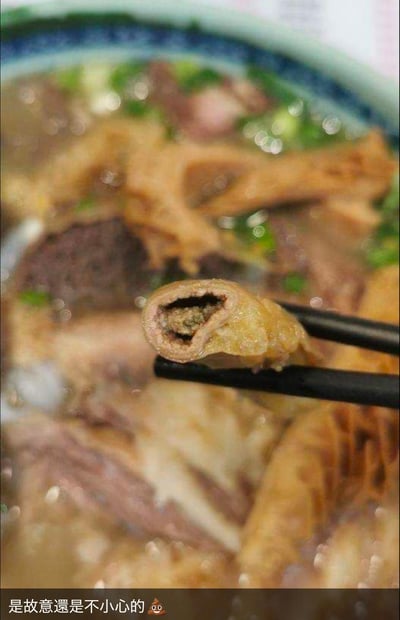 这是一张实拍的餐品照片，画面主体是一碗包含肥肠、血块等食材的汤类食物，筷子夹起一段肥肠，能清晰看到肥肠内部未清理干净，残留着污物。图片底部配有文字“是故意還是不小心的💩”，直白表达了就餐者吃到不干净食材的无语、嫌弃与吐槽，没有双关或谐音梗，核心是对餐厅餐品卫生状况的不满。