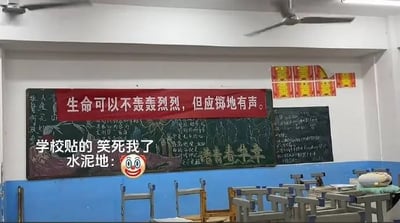 教室场景图，蓝色下墙白色上墙，天花板有吊扇。黑板上方悬挂红色横幅，写有标语“生命可以不轰轰烈烈，但应掷地有声。”。图片叠加文字“学校贴的 笑死我了 水泥地：🤡”，通过“掷地有声”与教室水泥地面的双关，制造幽默效果，调侃标语与实际环境的反差。