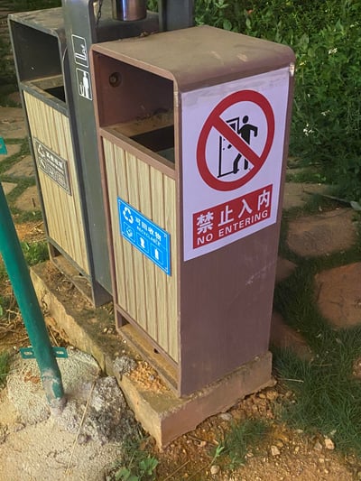 图片中展示了一个户外垃圾桶，主体为棕色金属材质，侧面贴有蓝色的“可回收物”分类标签。而在垃圾桶正面，却被人醒目地粘贴了一张“禁止入内”的标识，图案是一个人进入门口被红色圆圈斜线禁止，下方配有中英文“禁止入内 NO ENTERING”字样。
这一场景充满荒诞感——垃圾桶本是用于投放垃圾的公共设施，“禁止入内”的标识在此处显得完全多余，仿佛在一本正经地阻止人们“进入”垃圾桶，让人忍不住发笑。背景可见绿色植物和石板路，推测拍摄于公园或社区等户外公共区域。
