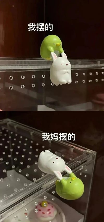 图片为上下对比结构，展示了同一组萌系玩偶的两种摆放方式。上半部分中，绿色小鸡玩偶在上、白色兔子玩偶在下，小鸡用爪子抓住兔子的耳朵，呈现出小鸡“拎起”兔子的活泼场景，上方配有文字“我摆的”；下半部分则相反，白色兔子玩偶在上、绿色小鸡玩偶在下，兔子用爪子抓住小鸡，形成兔子“拎起”小鸡的温馨画面，下方配有文字“我妈摆的”。两组场景均拍摄于透明展示盒内，背景为深色木质家具，整体风格可爱治愈，通过简单的摆放差异展现了家庭成员间不同的生活趣味。