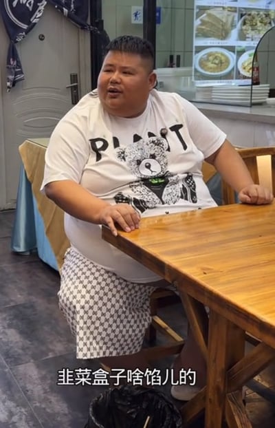 图片中是一位男子坐在餐馆的木质餐桌旁，他穿着印有泰迪熊图案和“PLANT”字样的白色T恤，搭配黑白格子短裤。背景可见餐馆的门（挂着深色布帘）、柜台以及展示的菜品图片。男子双手搭在桌沿，似乎正在询问。图片底部有文字“韭菜盒子啥馅儿的”，整体场景展现了日常餐馆中的一个普通瞬间。