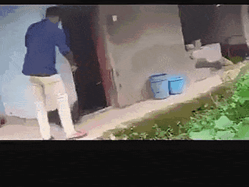 这是一段搞笑的动态GIF，画面呈现乡村房屋前的场景：一名身着蓝色上衣、白色长裤的男子，因地面与水面的视觉衔接形成错位，误将前方的水面认作平地，一脚踩上去后失去平衡滑倒，制造出滑稽的乌龙效果，利用视觉错觉带来笑点。