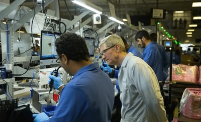 这张图片呈现了苹果公司前CEO蒂姆·库克身处现代化电子产品制造工厂的场景，他身着浅灰色工作服，正俯身专注观察一位身穿蓝色工装、佩戴蓝色手套的产业工人操作专业组装设备进行作业。车间内还有其他身着同款蓝色工装的工人在各自岗位忙碌，周围摆放着电脑终端、精密组装仪器、待加工组件以及包装好的产品，整体环境整洁有序，充满了工业生产的忙碌氛围，库克的神情专注，显然在细致了解产品生产的具体流程与细节。