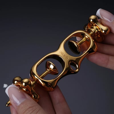 图片展示了一只手握着一个金色的OXO品牌工具，工具设计精致，表面光滑有光泽，带有两个圆形开口和可调节的金属部件。手部指甲修剪整齐，涂有白色指甲油，背景为深色，突出展示了工具的金色外观和细节。