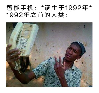 这是一张调侃时代科技发展差异的搞笑梗图，上方文字标注“智能手机：*诞生于1992年* 1992年之前的人类：”，引出下方的趣味画面。图中一位男子将老式固定座机举到脸前，模仿现代人们使用智能手机自拍的姿势，同时对着镜头比出手势，用这种夸张的违和反差，幽默地展现出智能手机出现前，当时的通讯设备与现代自拍文化的巨大差异，以此制造笑料，调侃科技发展带来的生活方式转变。
