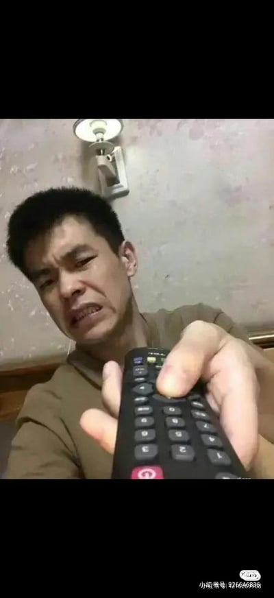 一名男子正手握黑色遥控器，眉头紧锁，嘴巴微张，表情看起来十分抓狂和无奈。背景是带有花纹的墙壁和一个白色的壁灯，整体场景像是在卧室里。