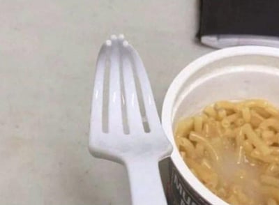 这张图片展示了一个存在严重生产失误的白色塑料叉子，它的叉齿并非正常叉子向下凹陷的结构，而是向上凸起的形态，完全无法实现叉子叉取食物的基本功能。叉子旁边放置着一杯泡好的通心粉杯面，这种完全不实用的餐具和食物的搭配，显得格外滑稽，让人对产品的品控问题感到哭笑不得。